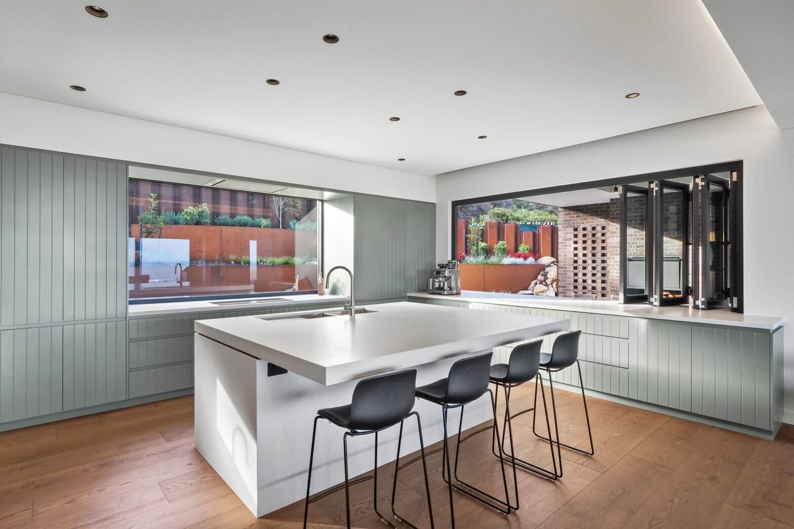 Modern open plan kitchen with large white island, sage green cabinetry, timber floors and floor-to-ceiling windows overlooking an alfresco area.